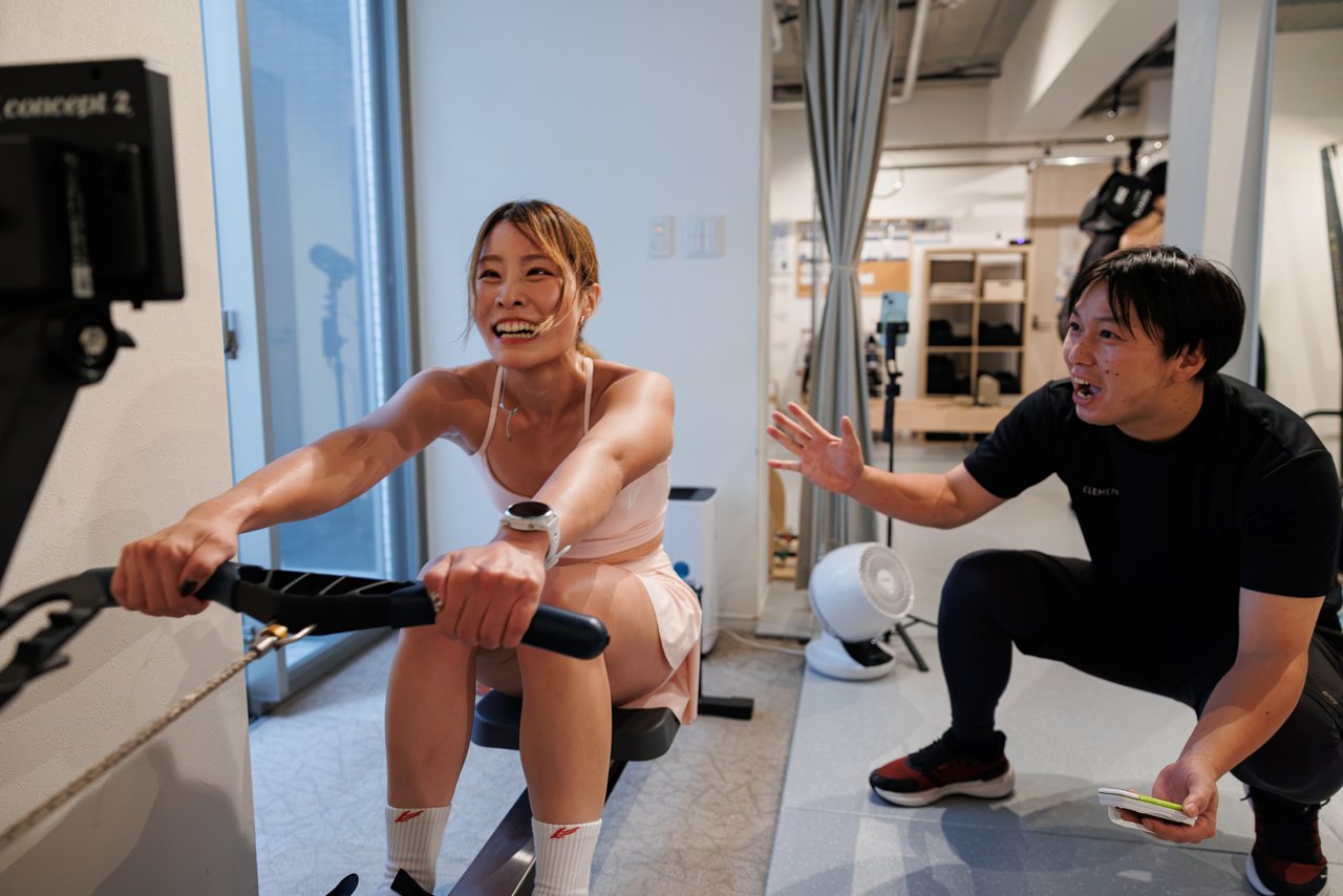ローイングマシンに取り組む女性と、声をかけるトレーナー